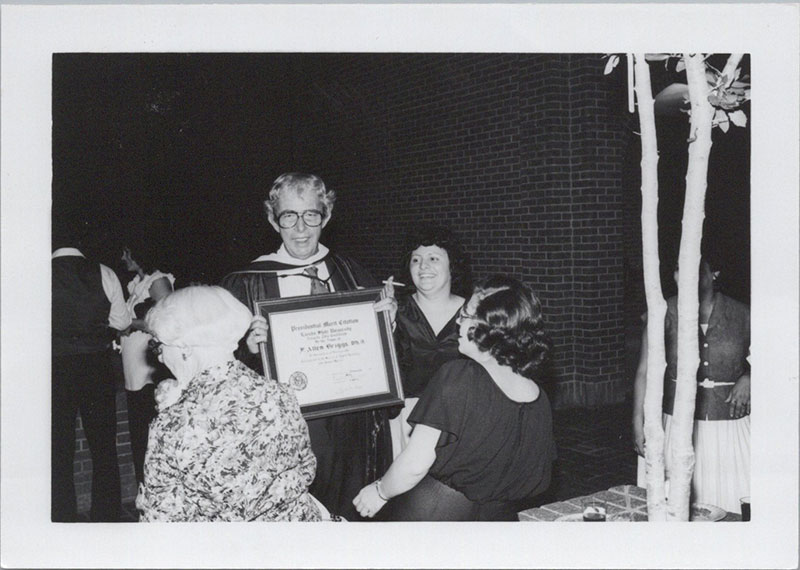 A smiling Dr. Briggs holds a certificate to the camera while being surrounds by friends.