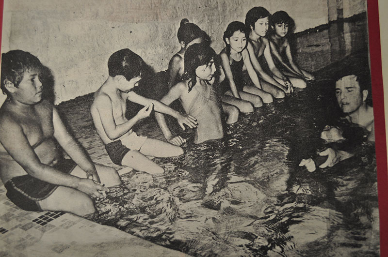 In a community pool, Dr. Ball teaches a young boy how to perfect his back stroke. Seven young children are sitting attentively on the pool's edge.